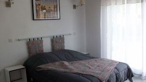 Un dormitorio con una cama grande, dos mesitas de noche, una mesita auxiliar, un cuadro en la pared y una ventana con cortinas.