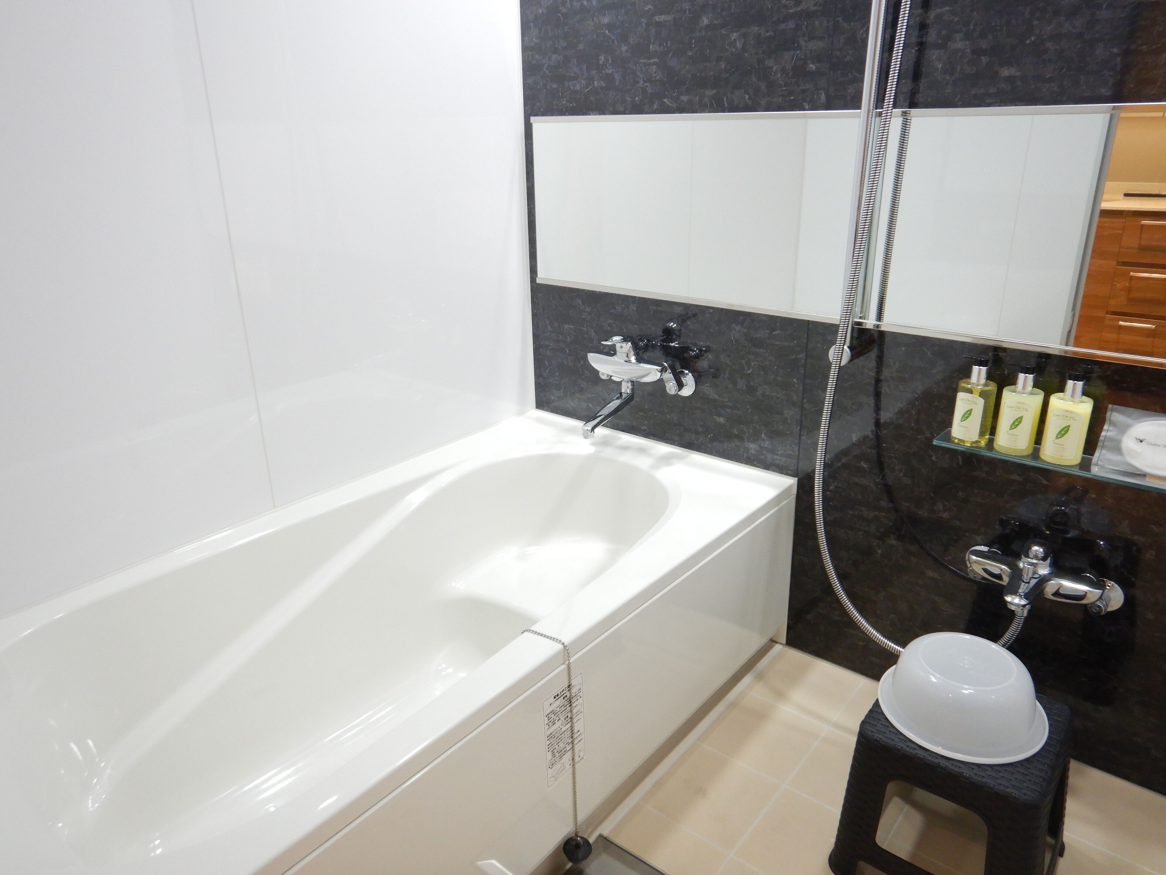 A modern bathroom with a white bathtub, a black and white tiled wall, a glass shelf with toiletries, and a small stool.