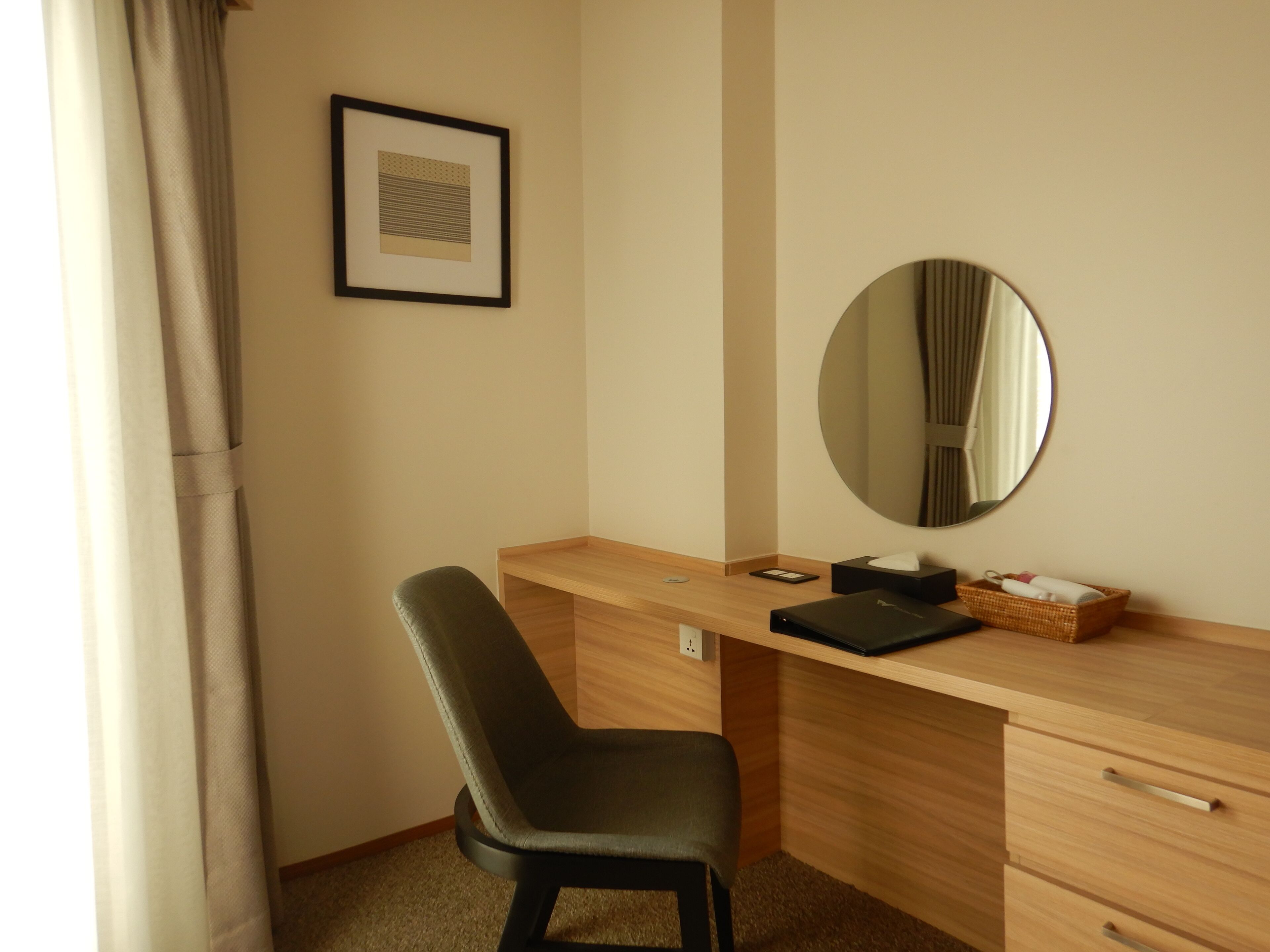 A hotel room with a wooden desk, a black chair, a round mirror, a framed picture on the wall, and a small basket with toiletries.