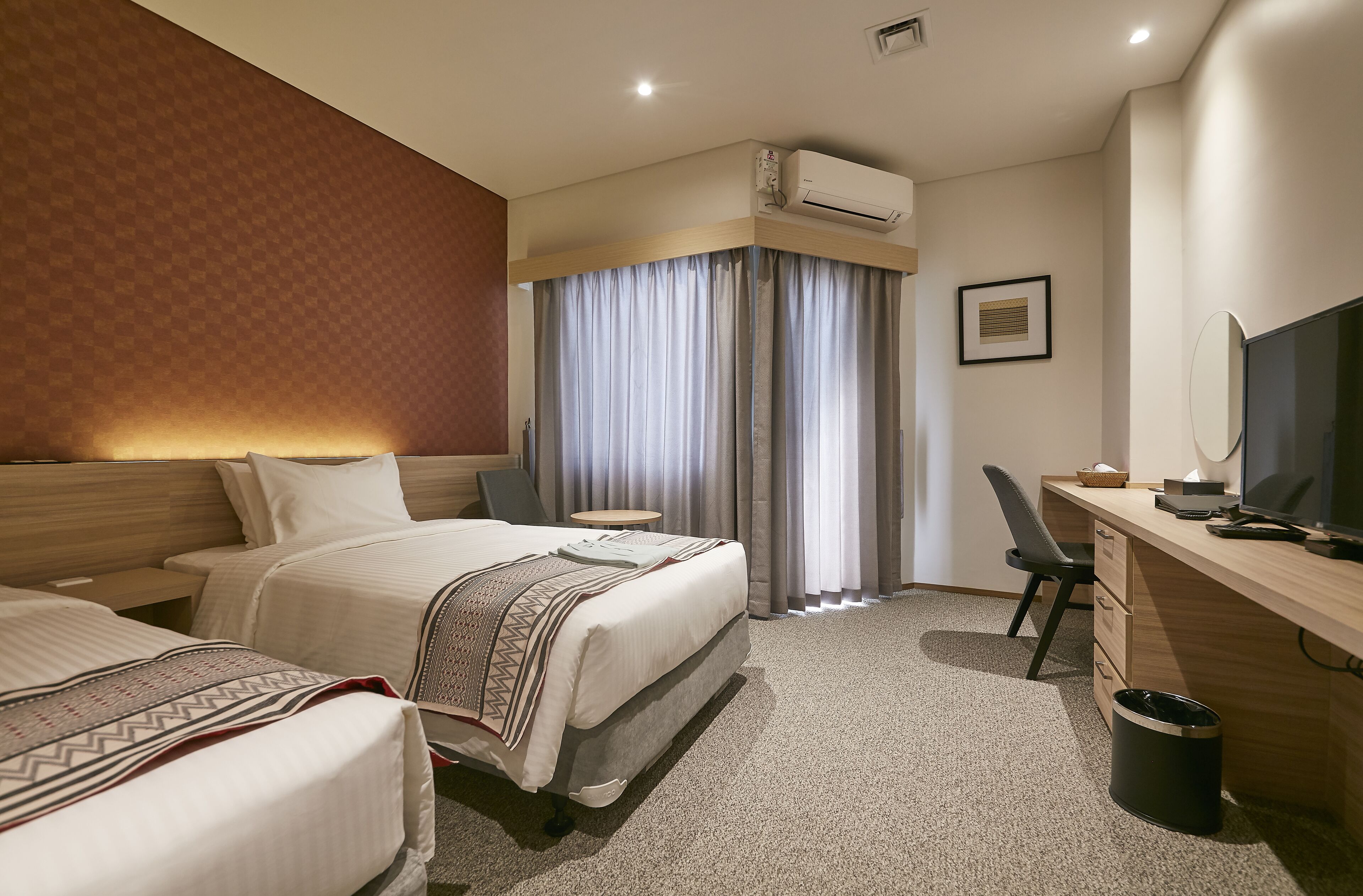 A hotel room with two beds, a desk with a computer, a TV, and an air conditioning unit.