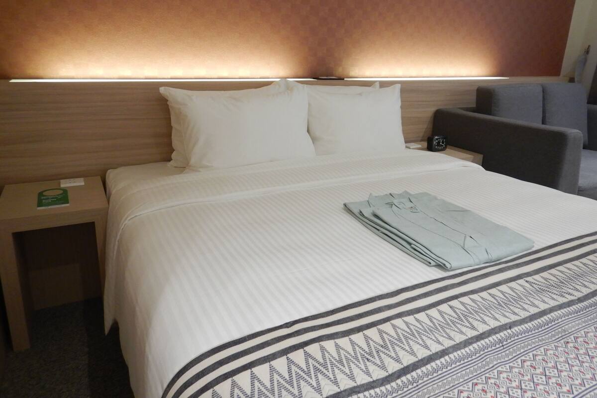 A hotel room with a neatly made bed, a bedside table with a green card, and a modern headboard with built-in lighting.