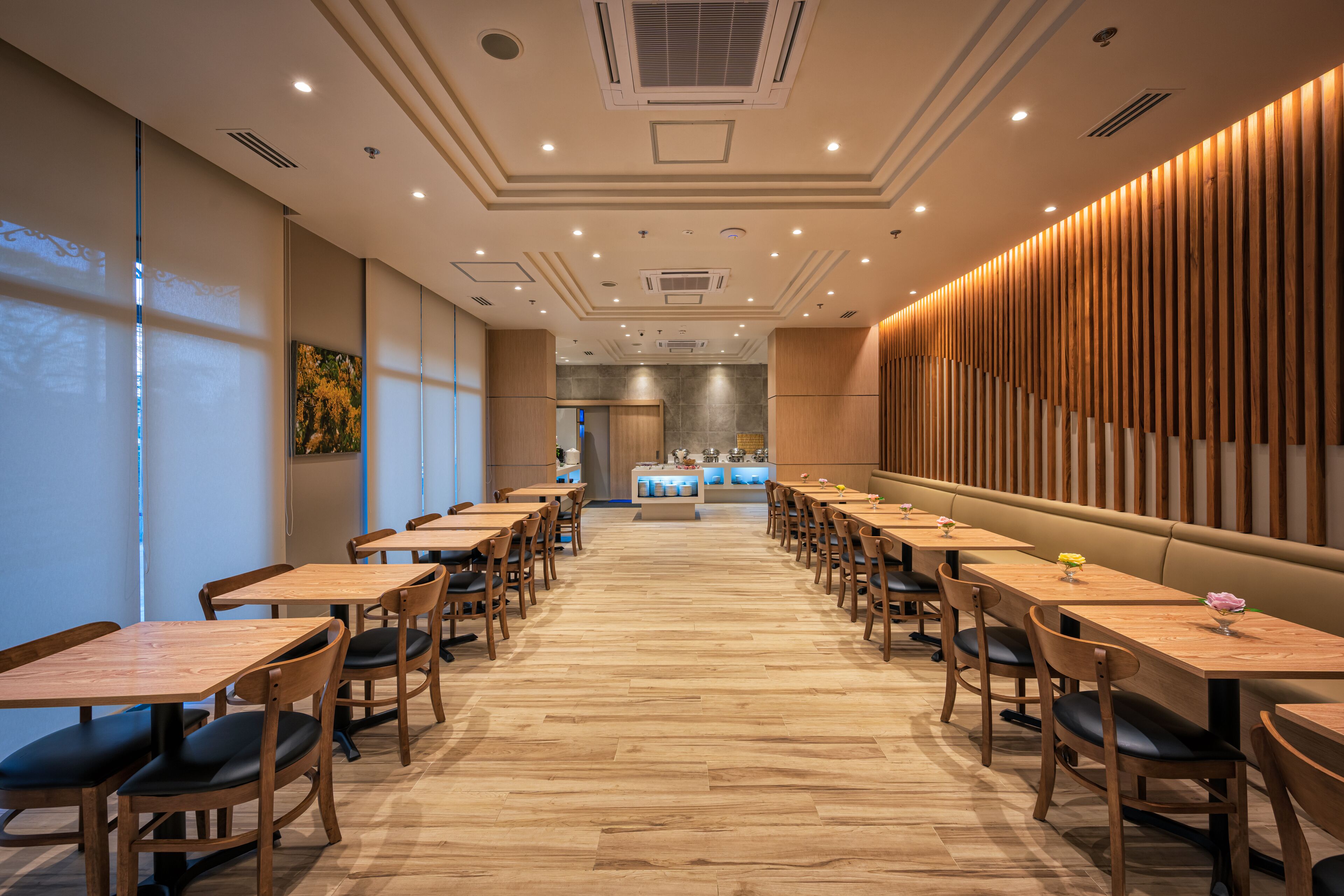 A spacious hotel dining area with multiple wooden tables and chairs, a central counter, and a modern ceiling design.