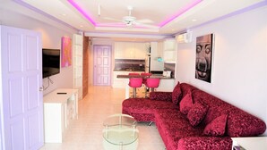 Un salon moderne avec un canapé rouge, une table basse en verre et un ventilateur de plafond. On aperçoit à l’arrière-plan un coin cuisine avec un four à micro-ondes et un évier.