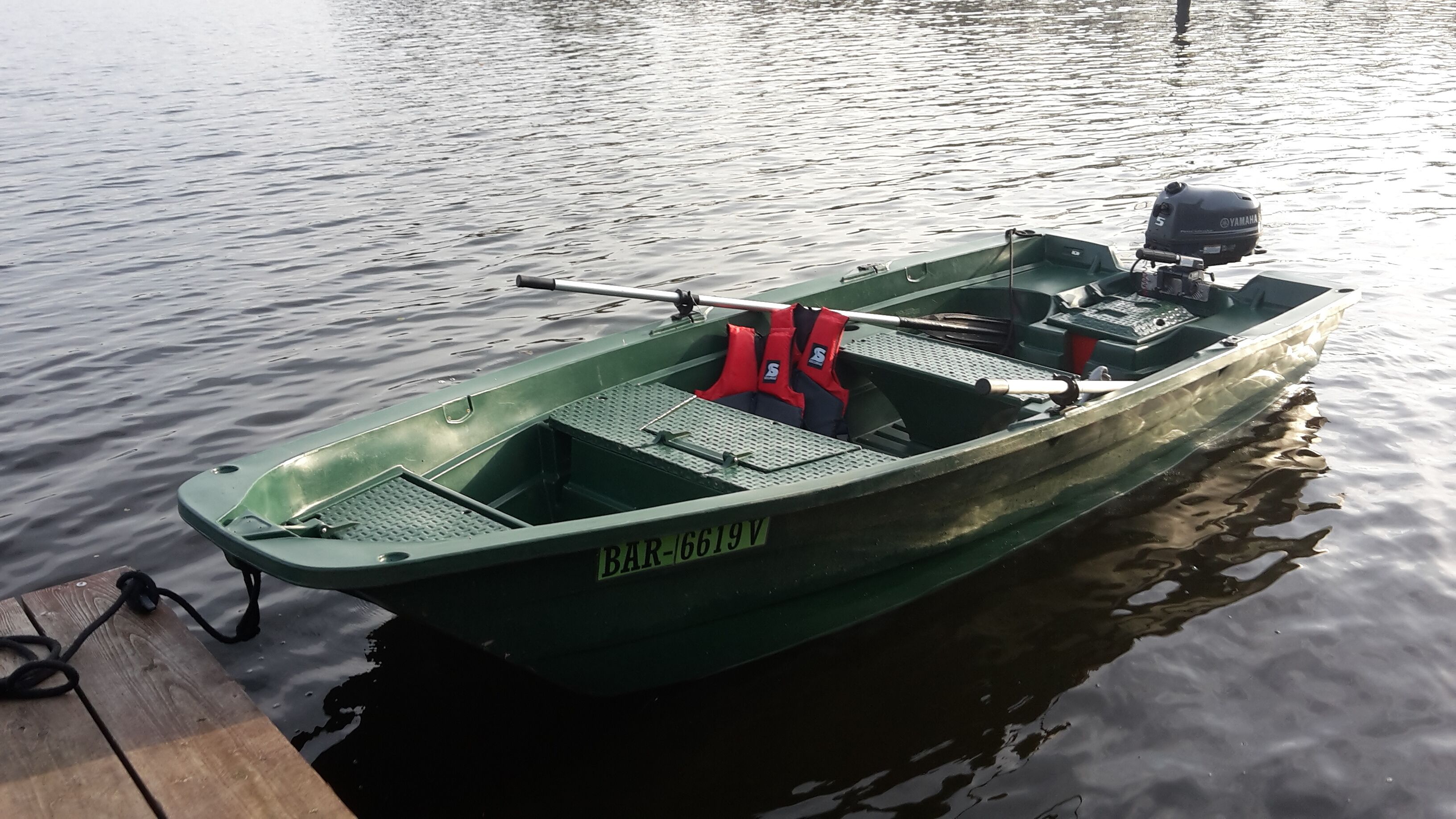 Un bateau à moteur vert, équipé d’un moteur hors-bord Yamaha, de gilets de sauvetage rouges et d’une canne à pêche, était amarré à un quai en bois.