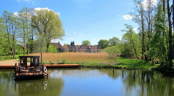 Un lago tranquilo con un muelle de madera y un bote pequeño, rodeado de abundante vegetación y una casa al fondo.