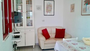 Une pièce avec un canapé beige, des coussins rouges, un meuble blanc aux portes vitrées, un téléviseur posé sur un meuble et une nappe à motifs floraux.