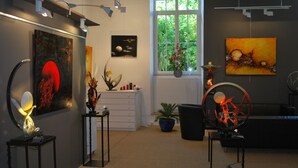 Een kunstgalerie met diverse kunstwerken die aan muren en tafels zijn tentoongesteld, een raam met uitzicht op groen, en een zwarte bank in de kamer.