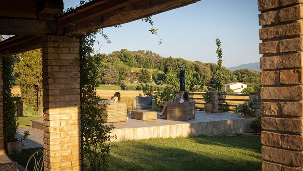 Un patio exterior con mobiliario de mimbre y una pared de piedra, con vistas a una zona verde y una colina.