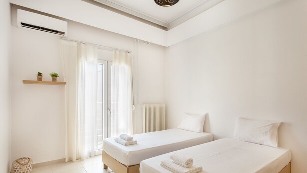 A hotel room with two beds, a window with curtains, a radiator, and a small shelf with plants. - Mirsini's Apartment in Chania Center (Chania)