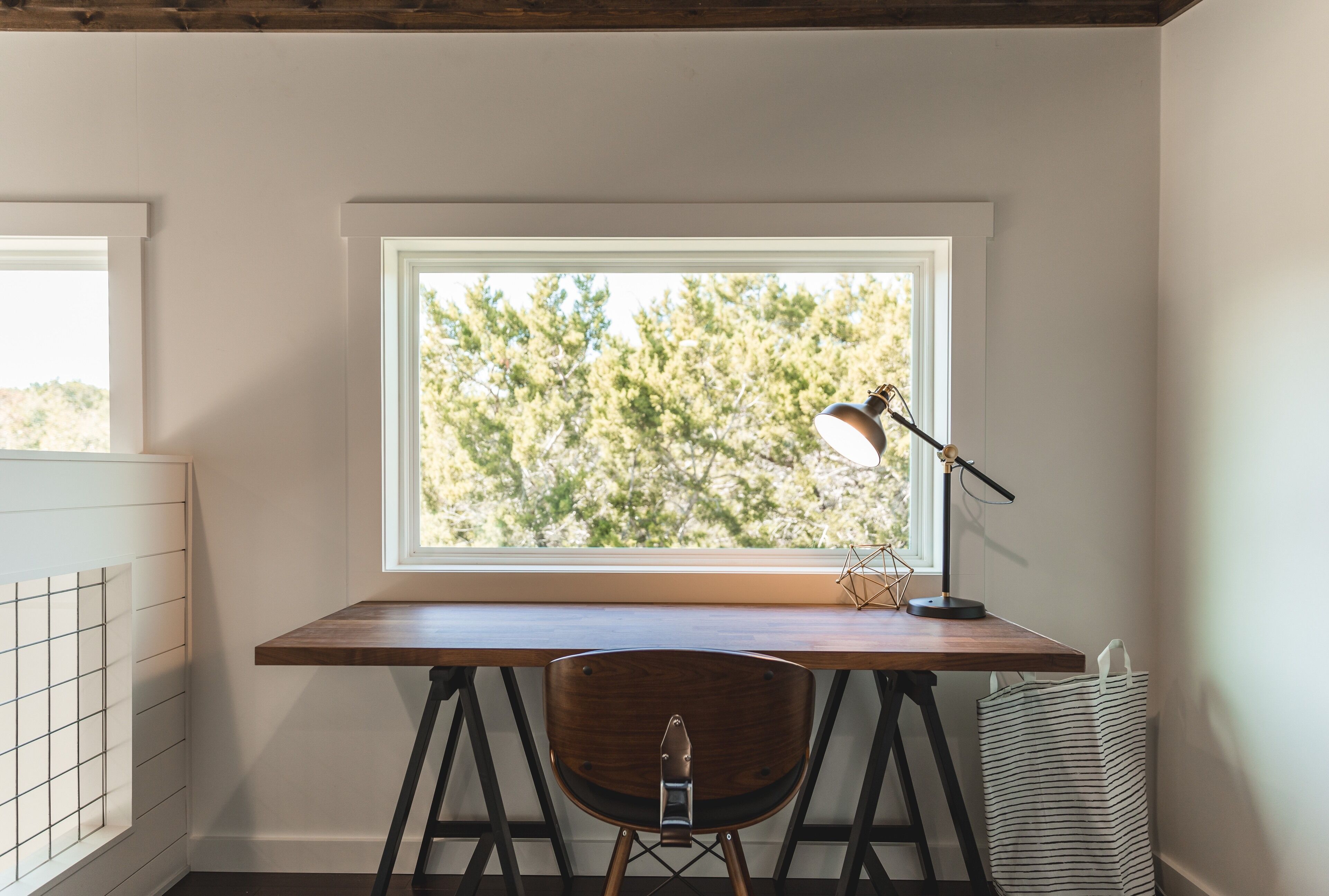 Een hoekwerkplek met een houten bureau, een stoel, een bureaulamp en een raam met uitzicht op bomen.