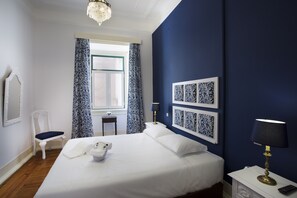 Un dormitorio ordenado con una cama grande, una mesita de noche con lámpara, una silla, una pequeña mesa y una ventana con cortinas estampadas.