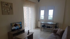 A living room with a TV, a sofa, a coffee table, a dining table with chairs, and a window with curtains. - Ria's Sea View Apartment (Chania)