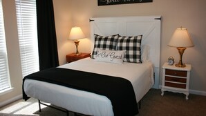 Una cama bien hecha con un cojín a cuadros blanco y negro y otro con la inscripción “Be Our Guest”. Hay dos lámparas de noche, una con un reloj en la mesita junto a la cama.