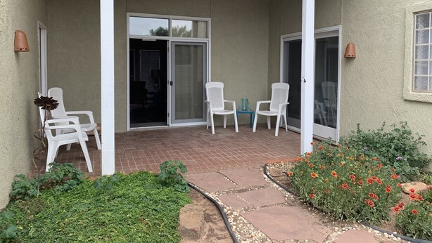 Un patio cubierto con sillas blancas, una puerta corrediza de vidrio y un jardín con flores y un sendero de piedra.
