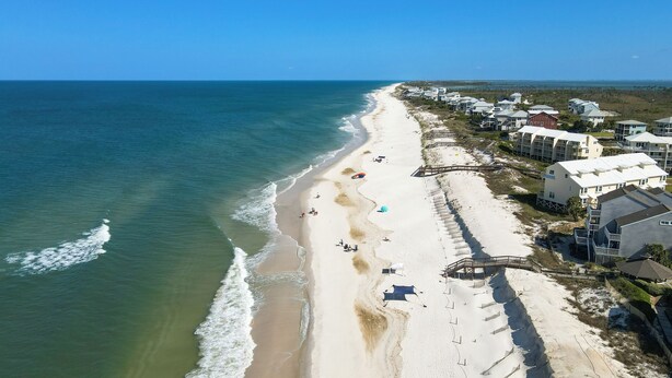 An aerial view of a beach with houses along the shore, a clear sky, and the ocean.