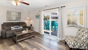 A living room with a sofa, coffee table, ceiling fan, and a view through a glass door to an outdoor area with chairs.