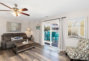 A living room with a sofa, coffee table, ceiling fan, and a view through a glass door to an outdoor area with chairs.