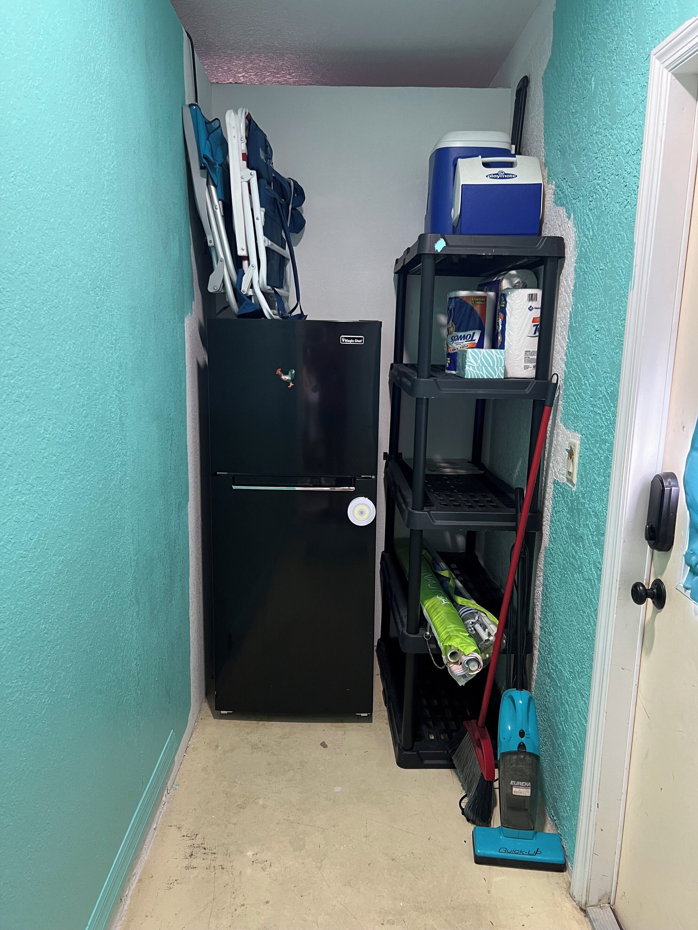 A compact kitchen with a black refrigerator, a blue and white Playmoto water dispenser, a broom, a vacuum cleaner, and a shelf with cleaning supplies.