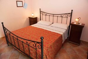 A single bed with a metal frame, a patterned bedspread, two pillows, a nightstand with a lamp, and a small cabinet. - Nel Giardino Dei Limoni (Montecorice)