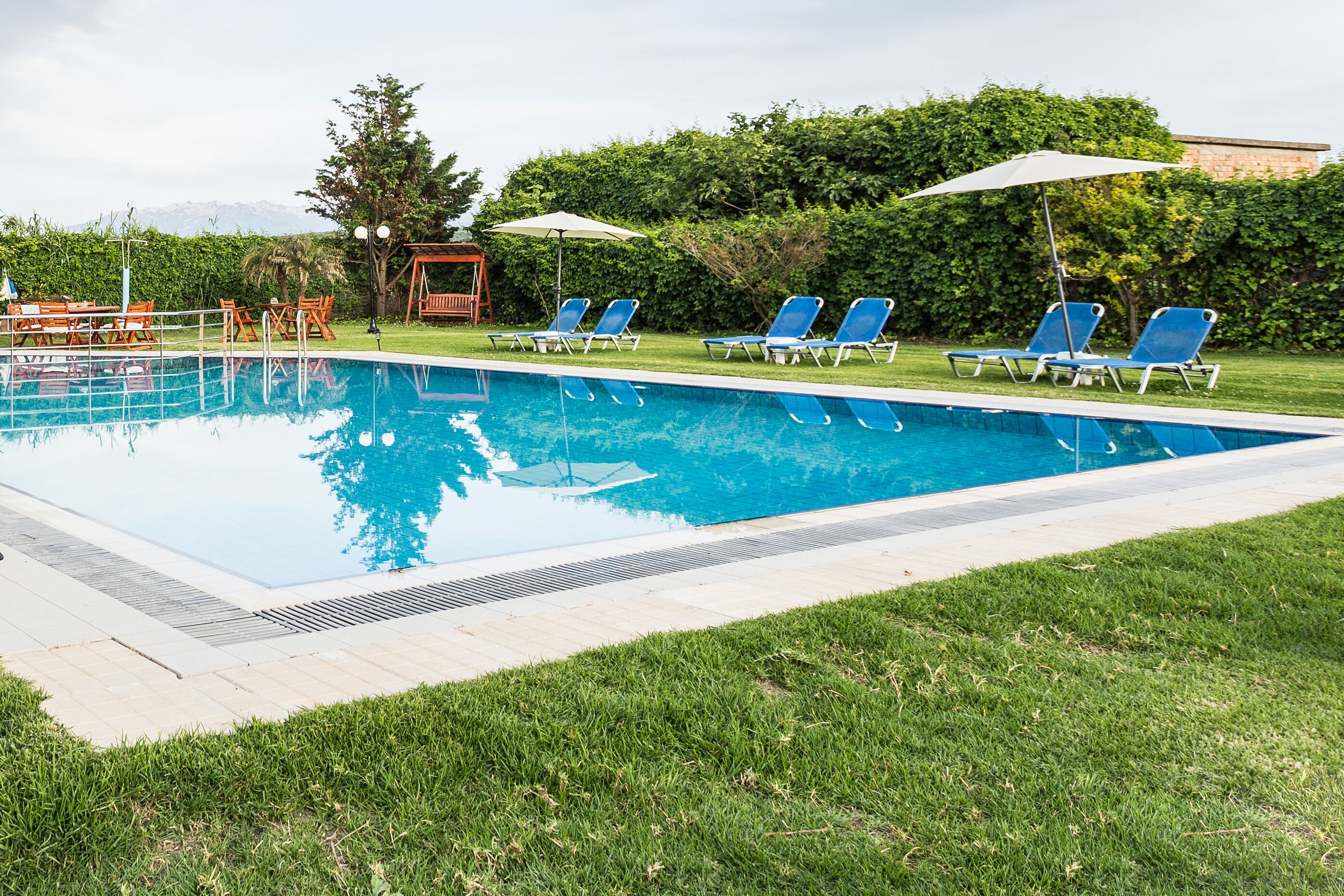 A rectangular swimming pool with a tiled edge, surrounded by a grassy area. There are several blue lounge chairs and umbrellas arranged around the pool.