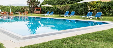 A rectangular swimming pool with a tiled edge, surrounded by a grassy area. There are several blue lounge chairs and umbrellas arranged around the pool.