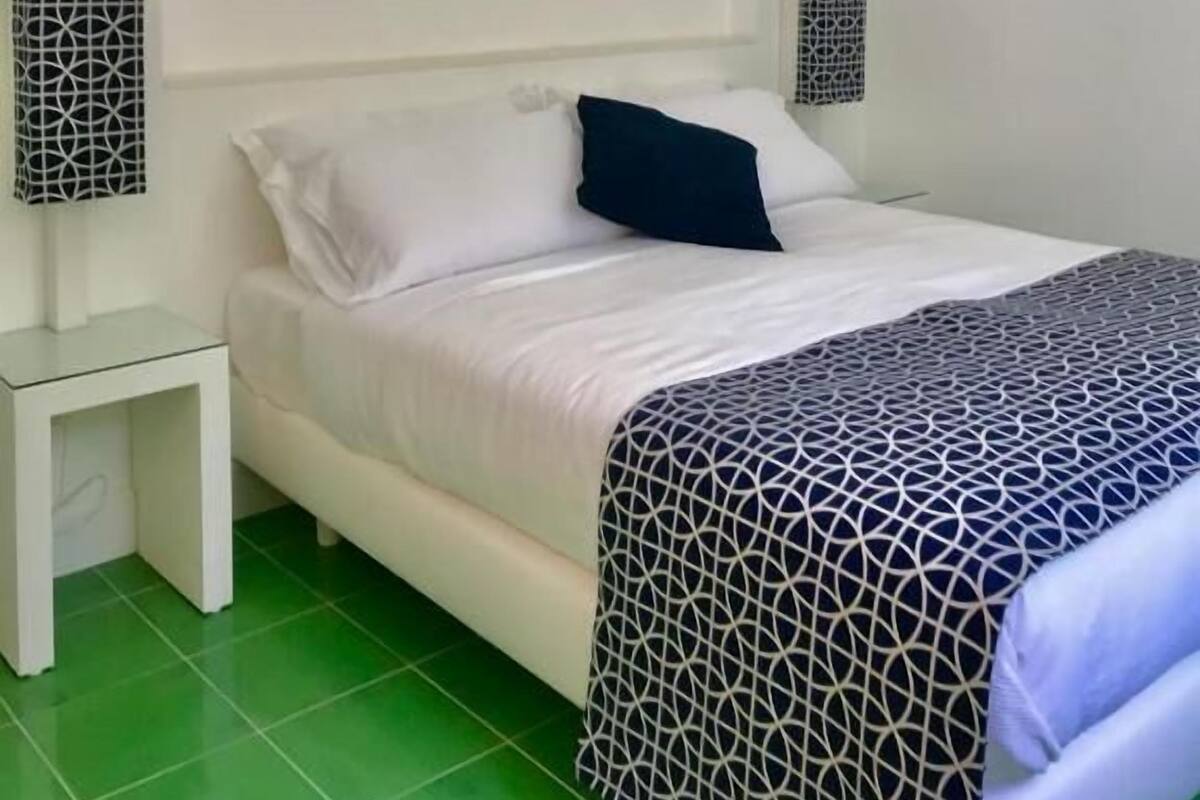 Une chambre à coucher avec un lit, un sol carrelé de couleur verte, des murs blancs et deux lampes fixées au mur.