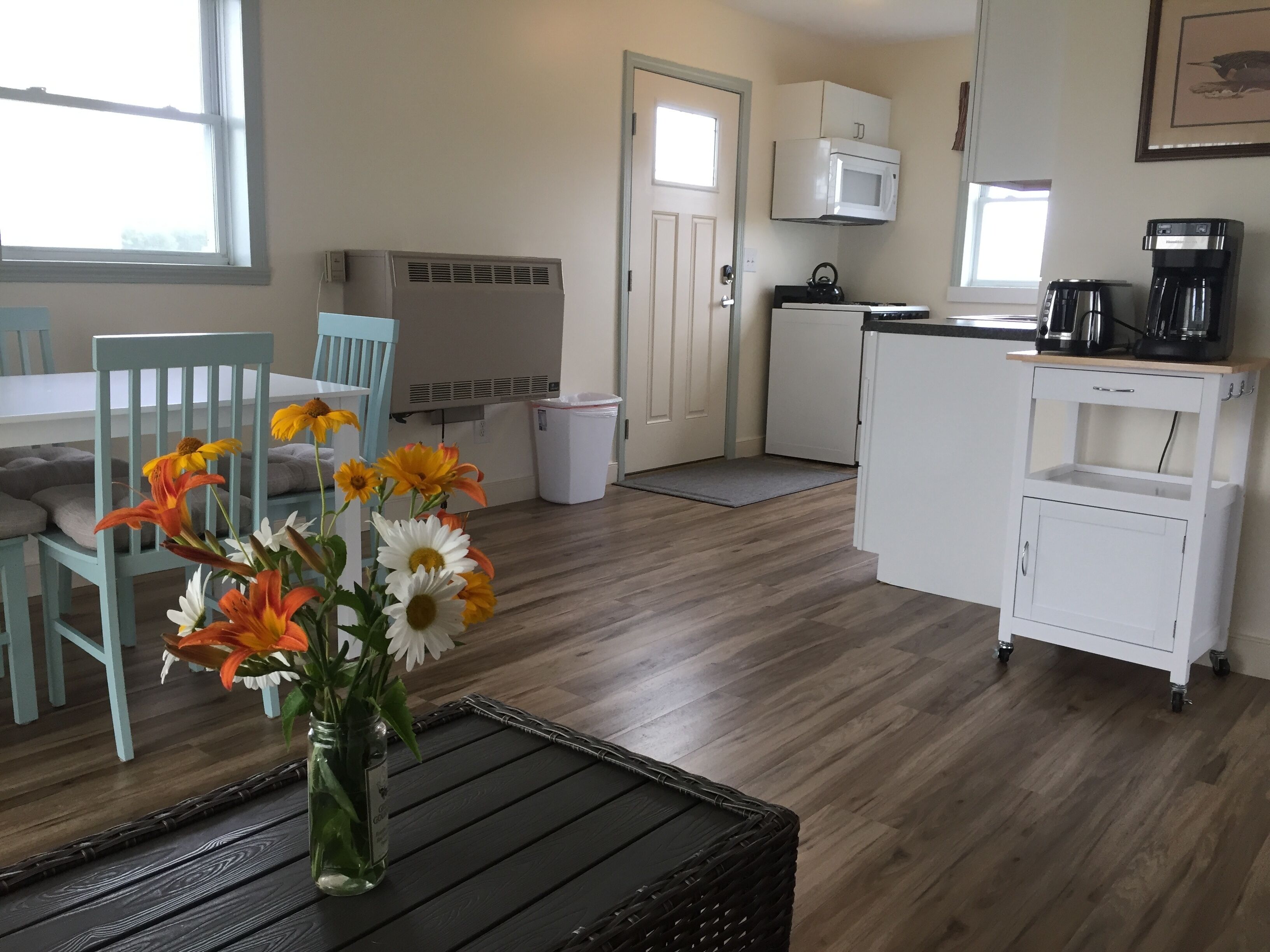Een compacte keuken met een magnetron, koffiezetapparaat en een kleine koelkast. Er is een eethoek met een tafel en stoelen, en een vaas met bloemen op een tafel.