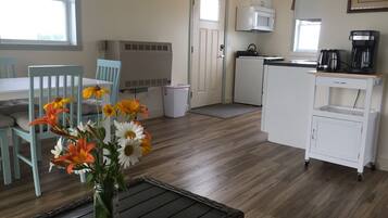 Een compacte keuken met een magnetron, koffiezetapparaat en een kleine koelkast. Er is een eethoek met een tafel en stoelen, en een vaas met bloemen op een tafel.