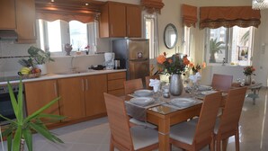 A modern kitchen with wooden cabinets, a stainless steel refrigerator, and a dining area with a glass table and wooden chairs. - Zoi's Holidays Apartment (Platanias)