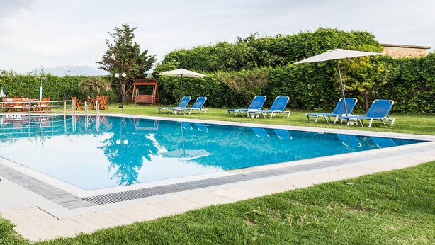 A rectangular swimming pool with a tiled edge, surrounded by a grassy area. There are several blue lounge chairs and umbrellas arranged around the pool. - Zoi's Holidays Apartment (Platanias)