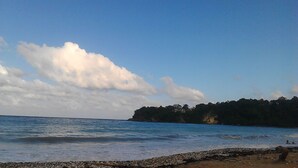 Una playa con aguas tranquilas, un litoral rocoso y un acantilado cubierto de bosque al fondo.