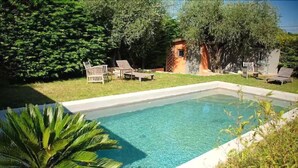 A rectangular swimming pool with clear blue water, surrounded by a white pool deck. There are lounge chairs and a small wooden structure in the background. - Le Clos des Bourges (Le Rouret)