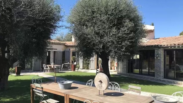 An outdoor dining area with a table and chairs, surrounded by olive trees and a building with a tiled roof. - Le Clos des Bourges (Le Rouret)