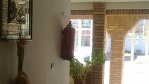 Un pasillo con un banco de madera, un jarrón con flores, un extintor y una planta grande en maceta.