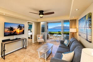 A hotel room with a TV, a sofa, a coffee table, a dining area, and a balcony with a view of the sea.