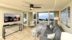 A spacious living room with a large flat-screen TV, a gray sofa, a coffee table, and a ceiling fan. - Chic and Remodeled Royal Mauian Beachfront  | Ground Floor Oceanfront Living (Kihei)