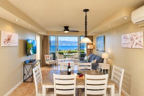 A spacious living area with a dining table, chairs, a sofa, and a television. There is a view of the ocean through large windows.