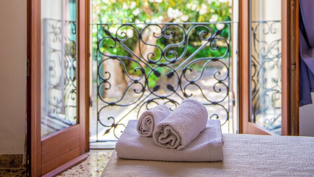 A room with a bed, a window with a view of greenery, and a balcony with a metal railing.