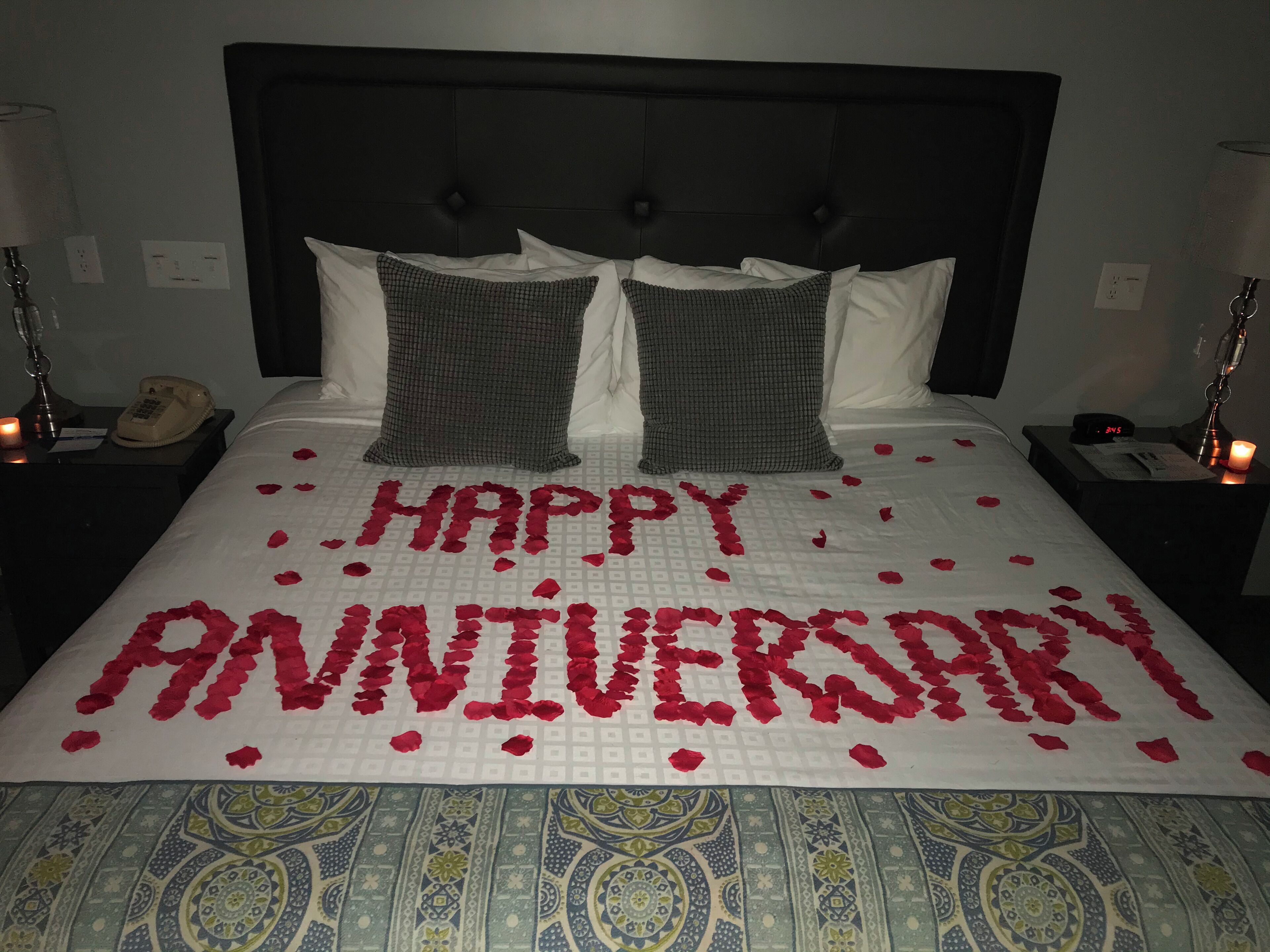 Uma cama com colcha branca e uma mensagem escrita em pétalas de rosa que diz "FELIZ ANIVERSÁRIO".