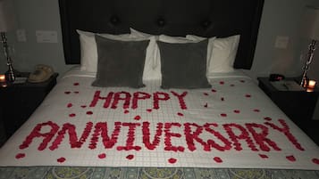 Uma cama com colcha branca e uma mensagem escrita em pétalas de rosa que diz "FELIZ ANIVERSÁRIO".
