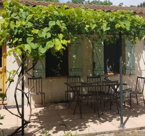 Um pátio coberto com mesa e cadeiras, uma pérgula com plantas trepadeiras e um edifício com persianas.