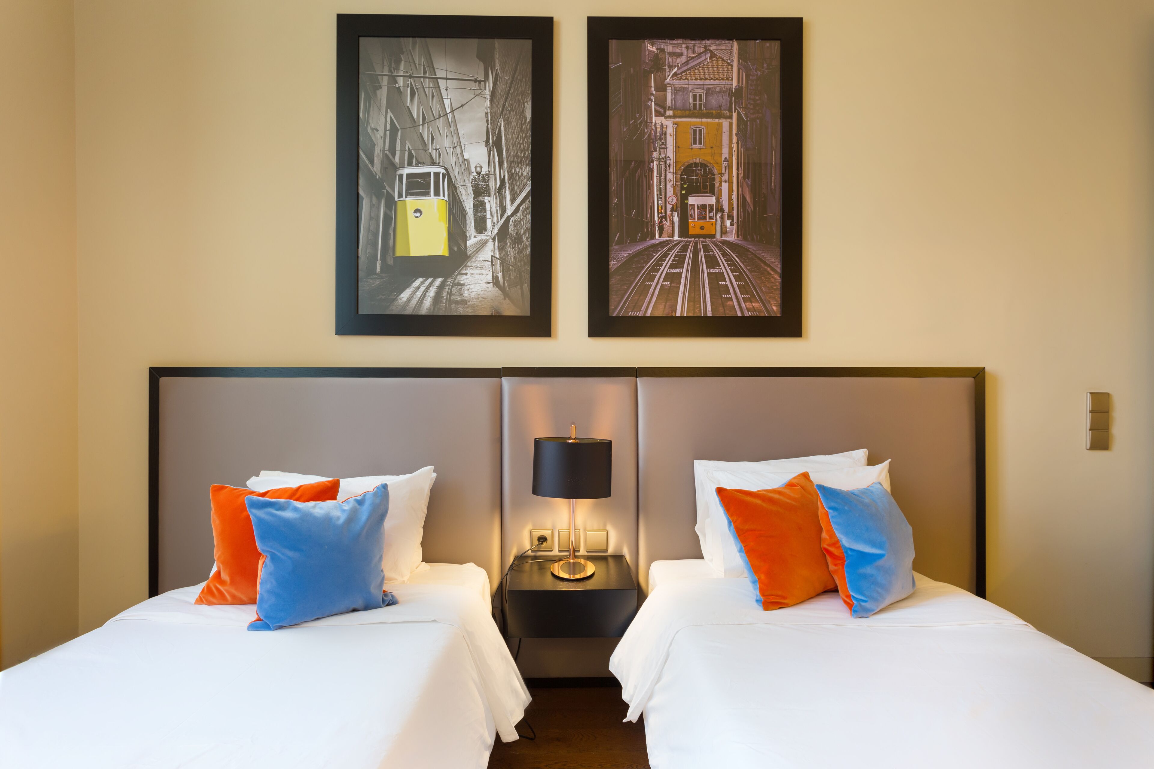 A hotel room with two beds, each with white bedding and orange and blue pillows. There is a nightstand with a lamp between the beds. Above the nightstand, there are two framed pictures of a tram on a street.