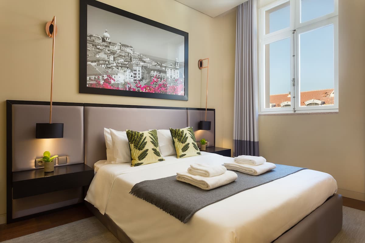 Ein ordentlich bezogenes Bett mit Kissen, ein Nachttisch mit Lampe und ein Stadtbild im Bilderrahmen an der Wand.