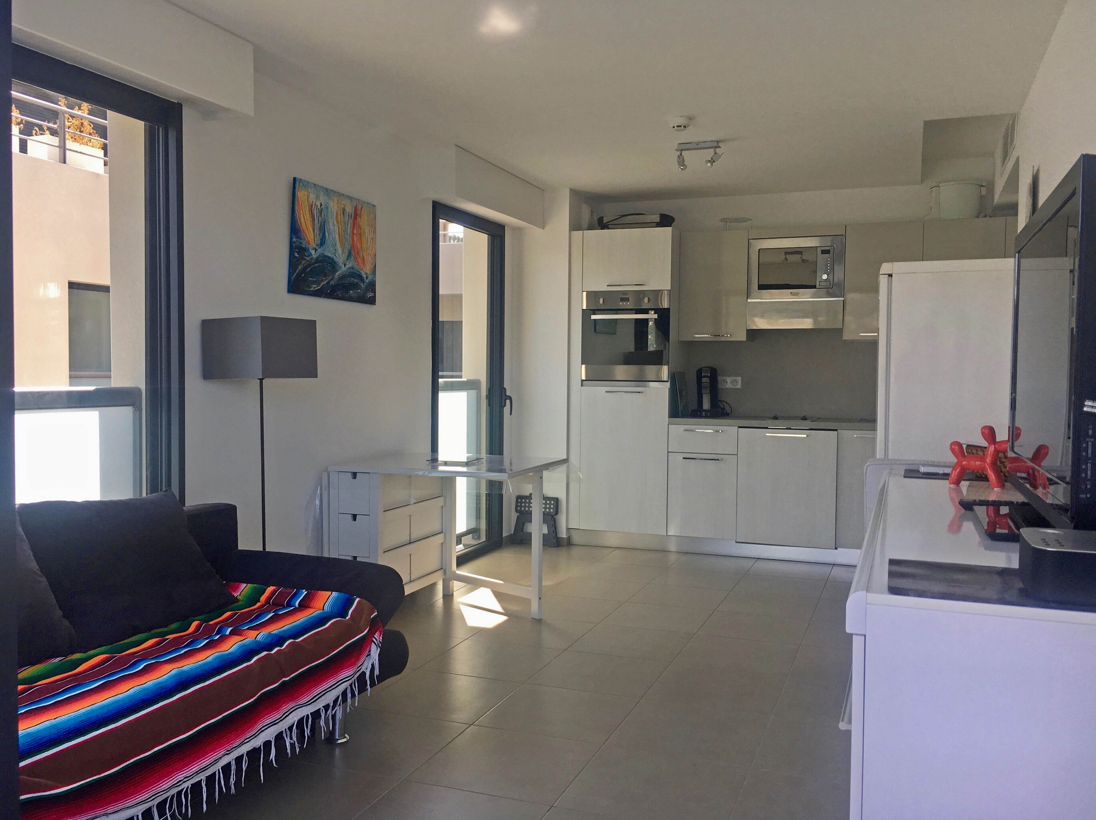 Una sala moderna con sofá, mesa de centro y manta colorida. El área de la cocina incluye un refrigerador, microondas y estufa.