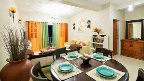 Un comedor con una mesa redonda dispuesta para cuatro personas, rodeada de sillas. Se observa una estantería con libros y objetos decorativos, un sofá y un aparador al fondo.
