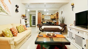 Una sala de estar con sofá, mesa de centro y televisión. Se observa una cocina al fondo con armarios de madera y una mesa de comedor.
