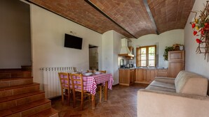 A spacious living area with a staircase, a dining table with chairs, a sofa, and a kitchen area with wooden cabinets and a window. - Apartment I Reucci Trilo5 (Pomarance)