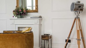A room with a white fireplace, a mirror with a gold frame, a tripod with a light, and a brown sofa. - My Opera de Paris II (Paris)