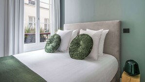 A neatly made bed with white linens and two green decorative pillows, a window with white curtains, and a green heater on a wooden stool. - My Opera de Paris II (Paris)