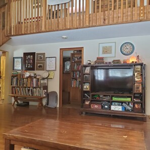 Amplio salón con suelo de madera, un televisor de pantalla plana grande y una estantería llena de libros y objetos decorativos.